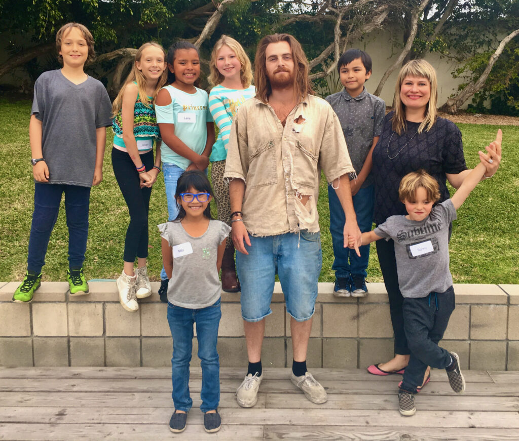 Mikey Mike with a children’s choir and Charissa Nielsen outdoors on a deck, trees and grass behind them.